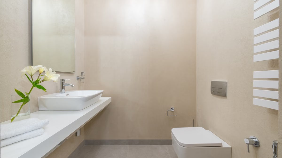 White ceramic toilet placed against wall with mirror and sink near blooming white flowers and towels in light modern bathroom